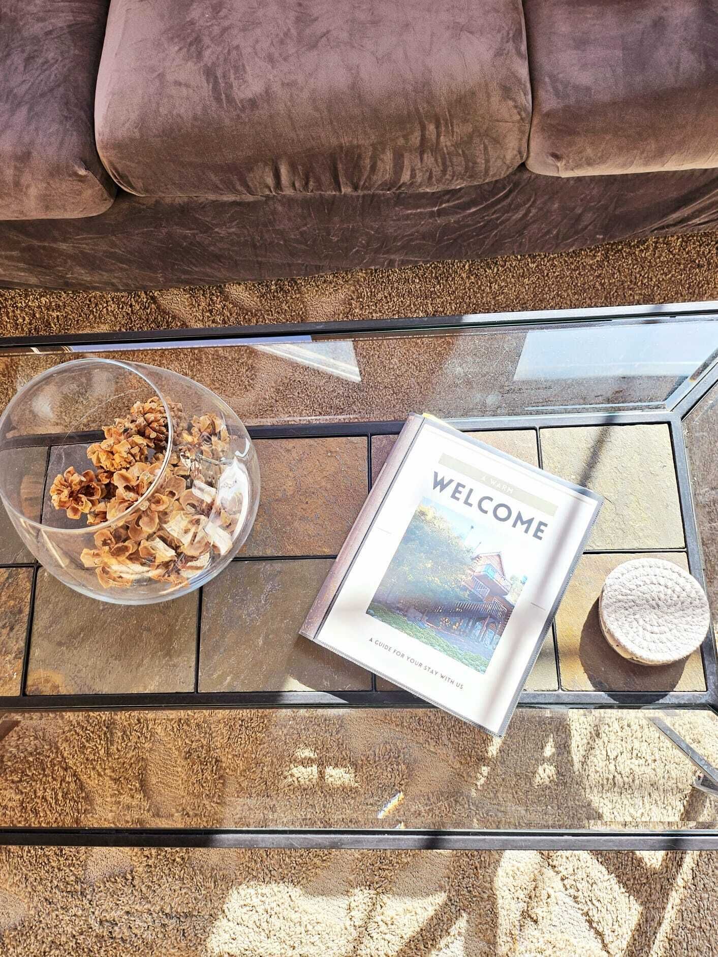 A glass-top table with a bowl of snacks, a welcome brochure, and a small round object.