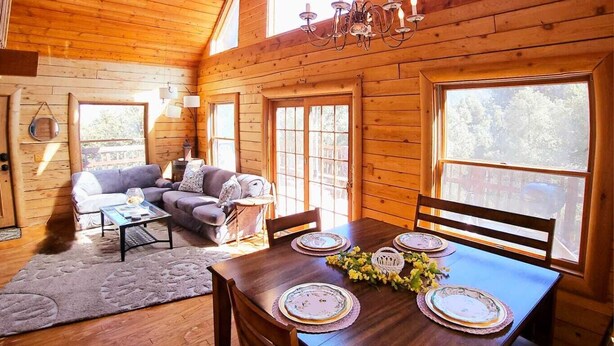 A wooden cabin interior with a dining table set for four, a sofa, and a ceiling fan.