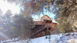 A wooden cabin with a staircase leading to a deck, surrounded by trees and snow.