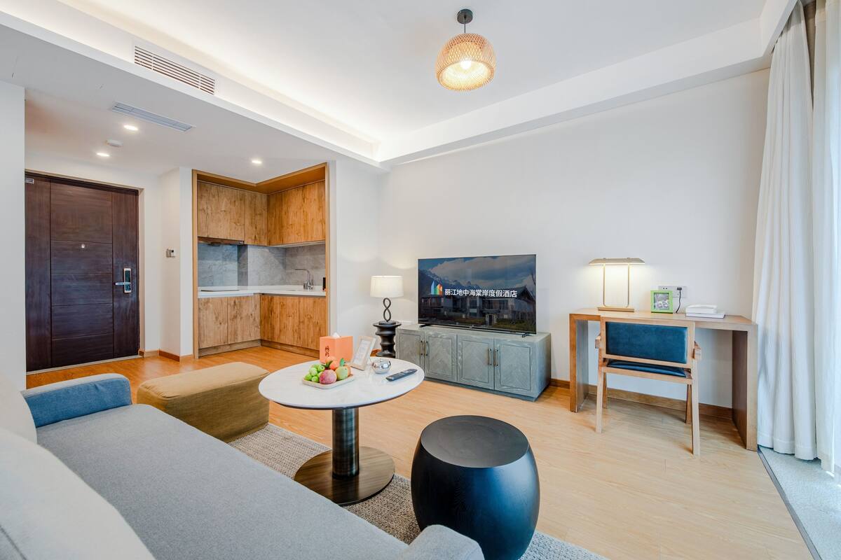 Una sala de estar moderna con un sofá, una mesa redonda de centro y un televisor. Cuenta con una zona de cocina con armarios de madera y una mesa de comedor.