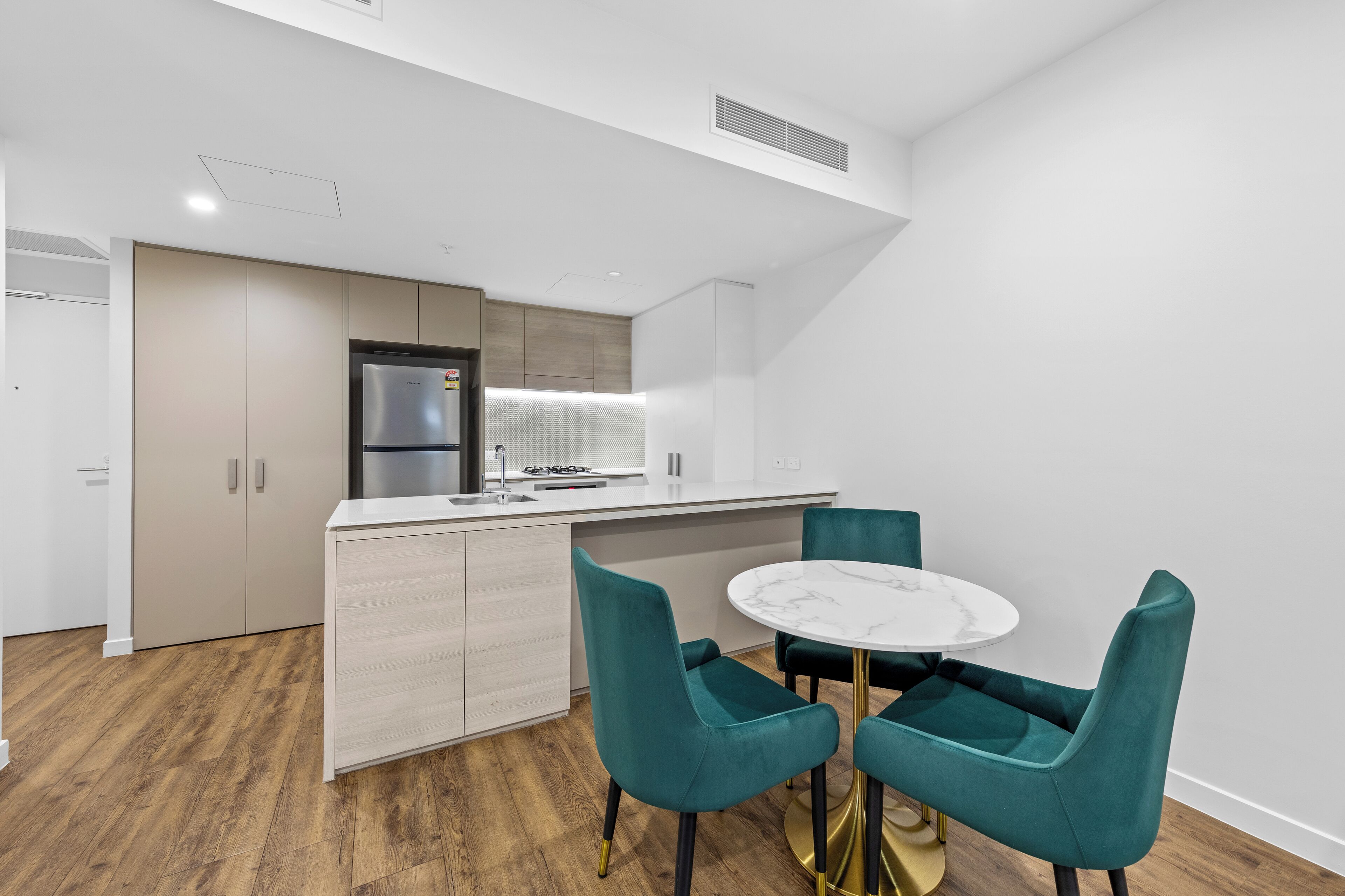 Eine moderne Küche mit integriertem Kühlschrank, einer Mikrowelle und einem Gasherd. Es gibt eine weiße Arbeitsplatte und ein Spülbecken. Der Boden ist aus Holz und die Schränke sind weiß. Vor der Küche steht ein runder Tisch mit zwei türkisfarbenen Stühlen.