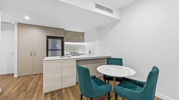 Eine moderne Küche mit integriertem Kühlschrank, einer Mikrowelle und einem Gasherd. Es gibt eine weiße Arbeitsplatte und ein Spülbecken. Der Boden ist aus Holz und die Schränke sind weiß. Vor der Küche steht ein runder Tisch mit zwei türkisfarbenen Stühlen.