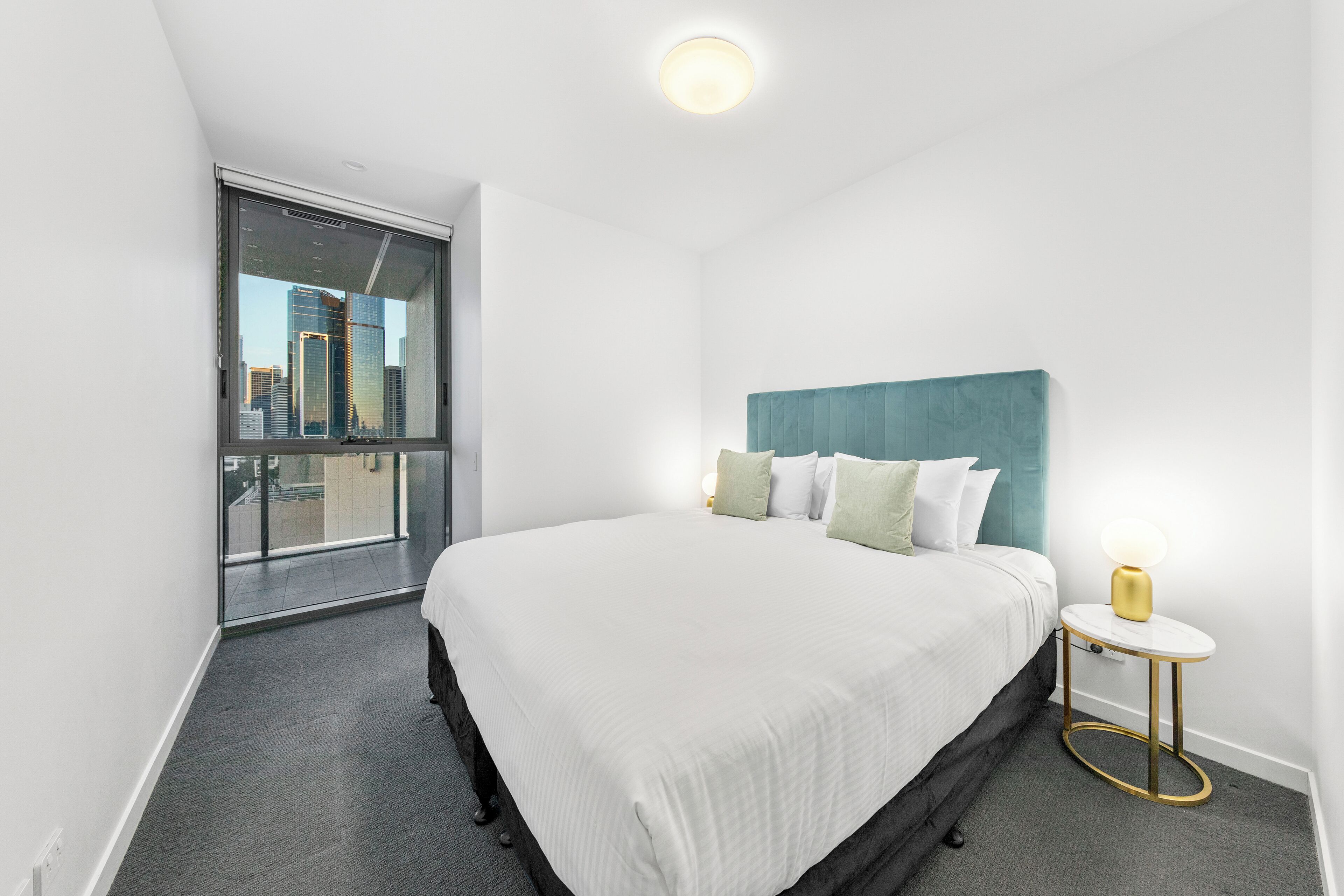 Una habitación de hotel moderna con una cama grande, vista de la ciudad a través de un ventanal y una mesita de noche con lámpara.