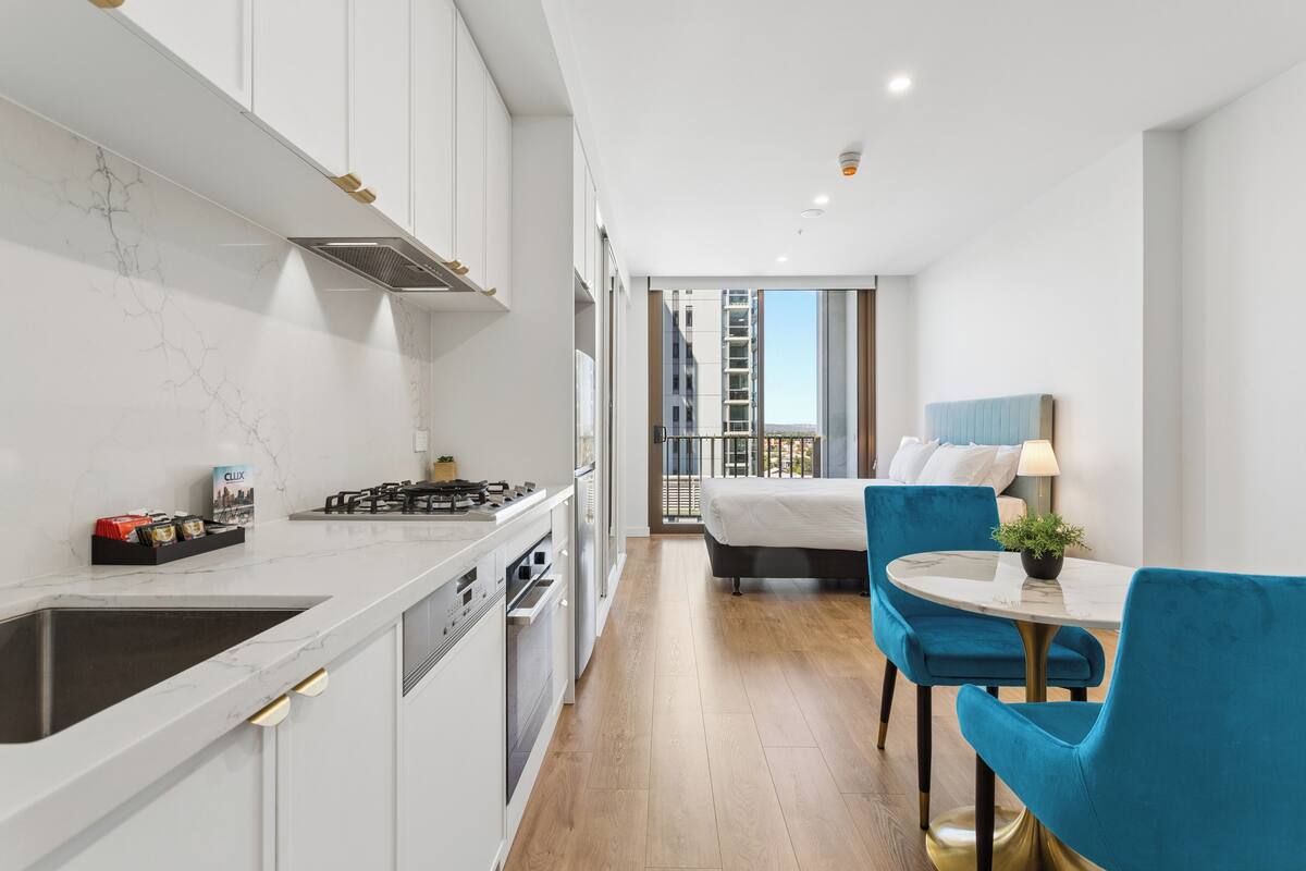 A modern kitchen with white cabinets, a marble backsplash, and a sink. There is a dining area with a round table and blue chairs. A bed with white bedding is visible in the background.
