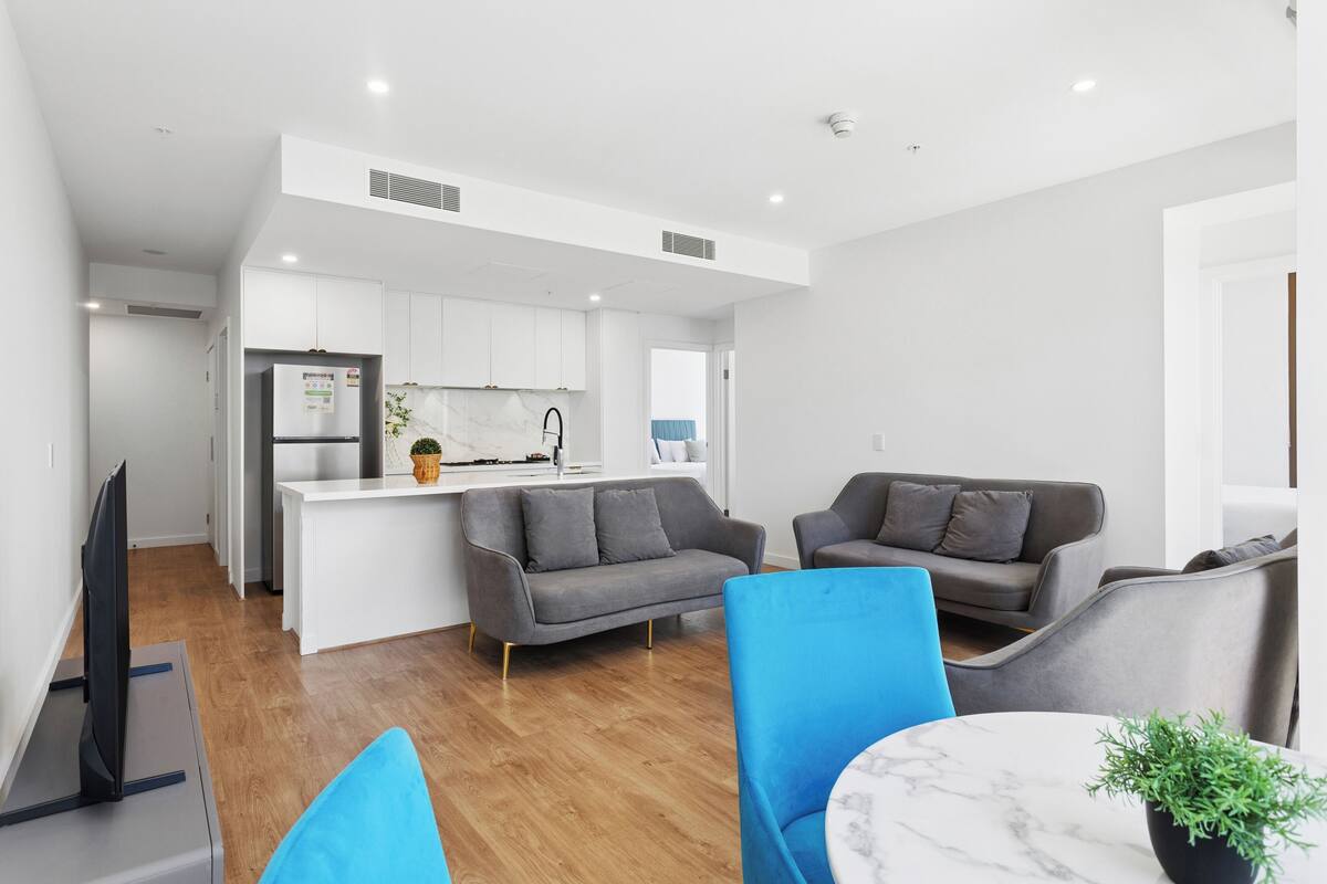 A modern living space with a kitchen, grey sofas, a blue armchair, and a marble table.