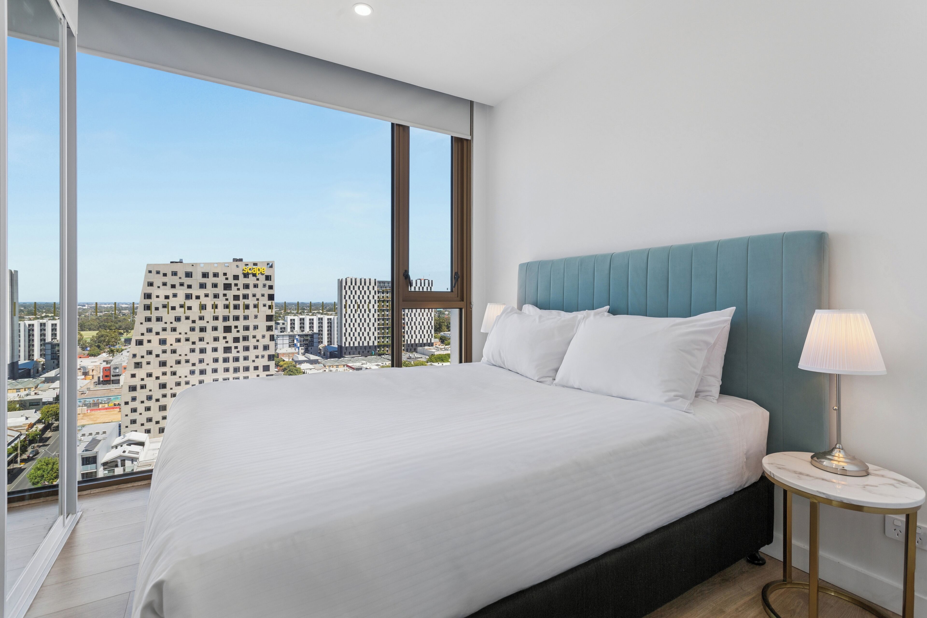 A hotel room with a large bed, a nightstand with a lamp, and a view of the cityscape through large windows.