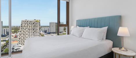 A hotel room with a large bed, a nightstand with a lamp, and a view of the cityscape through large windows.