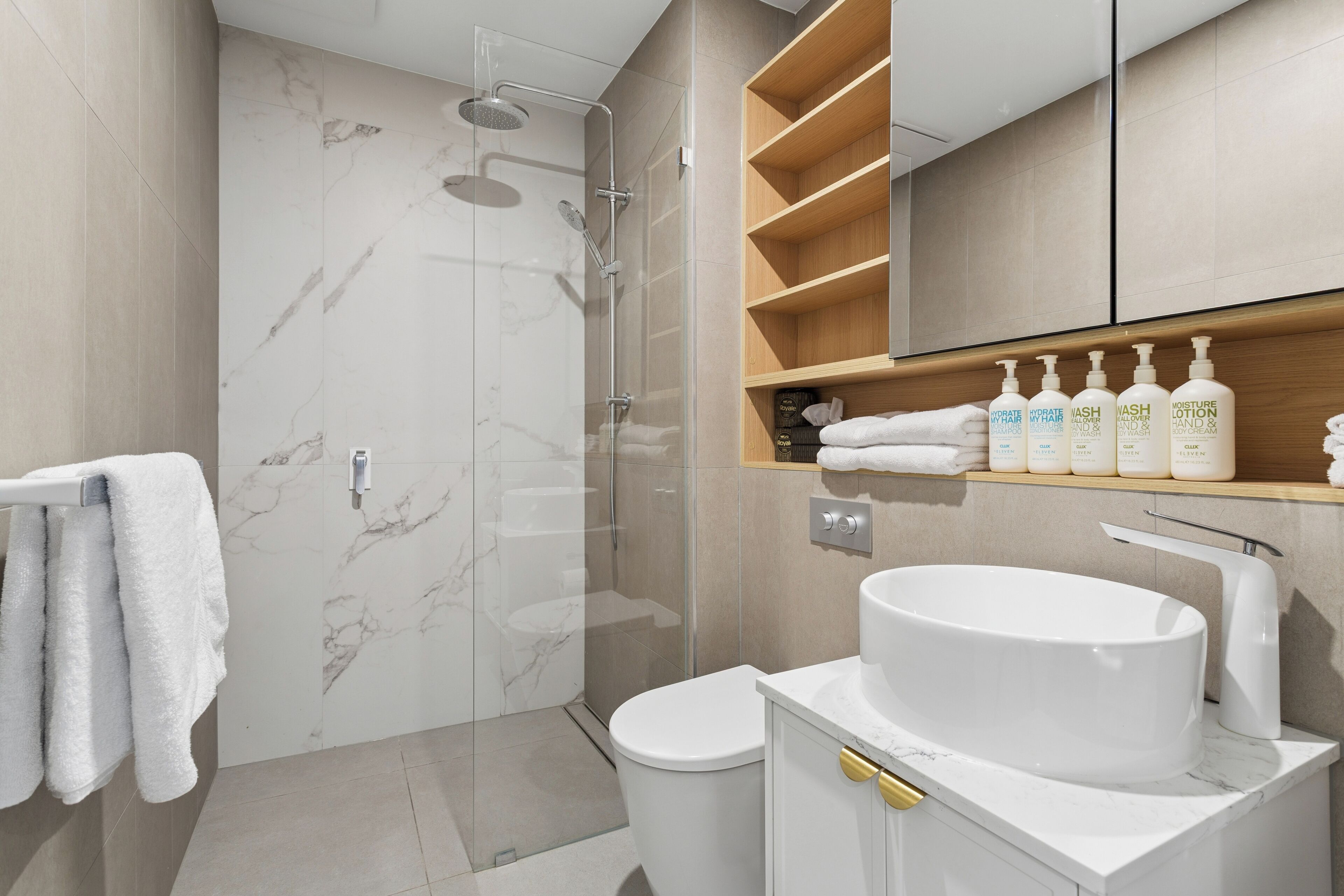 A modern bathroom with a glass-enclosed shower, a white sink, and a wooden vanity with toiletries.