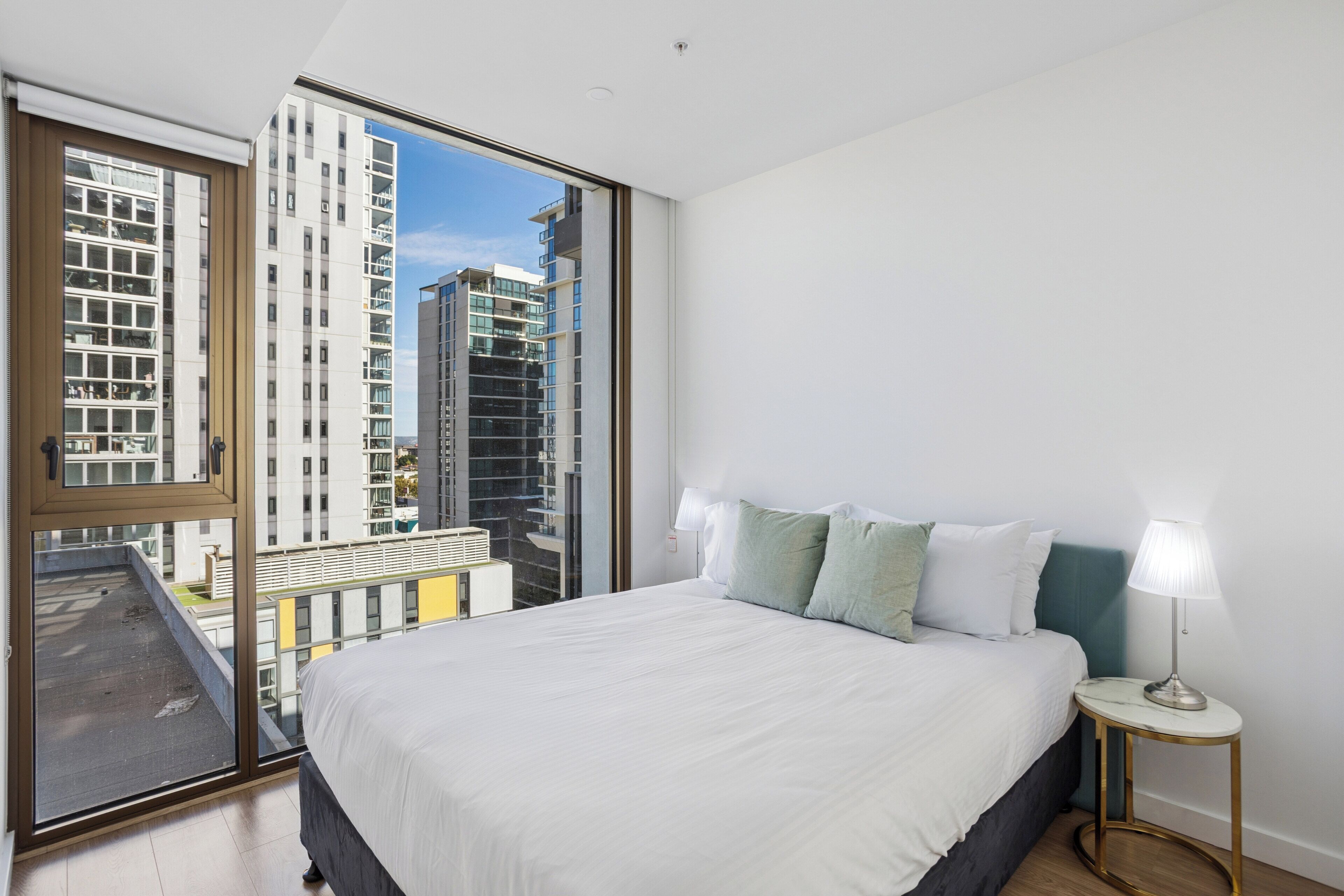 A modern hotel room with a large bed, a bedside table with a lamp, and a view of tall buildings through a large window.