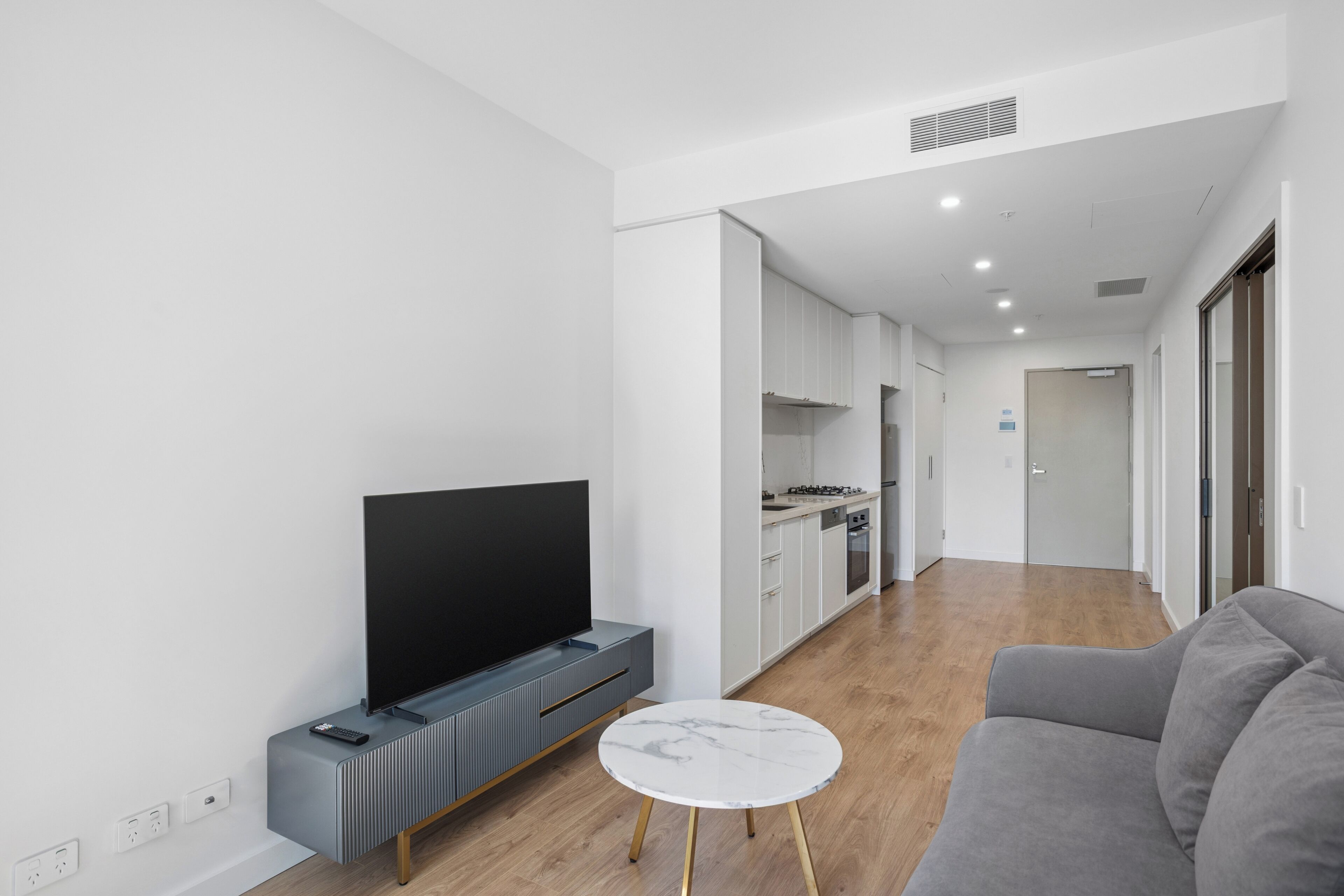 A modern living room with a flat-screen TV, a gray sofa, a small round coffee table, and a kitchen in the background.