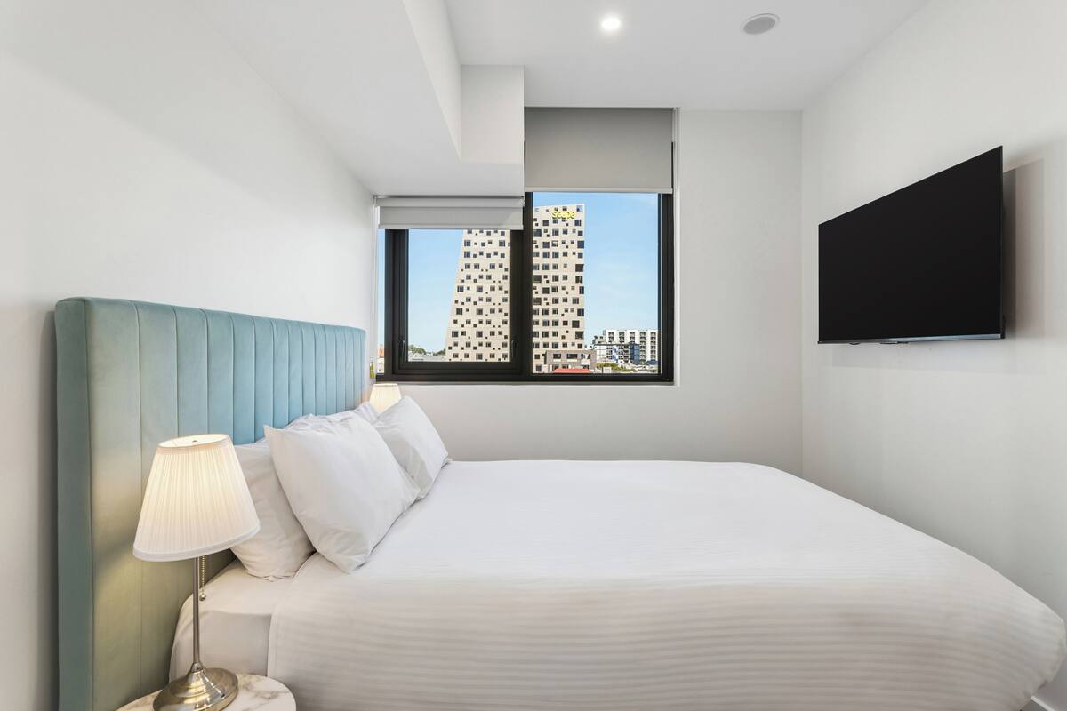 A hotel room with a bed, a nightstand with a lamp, a TV mounted on the wall, and a window with a cityscape view.