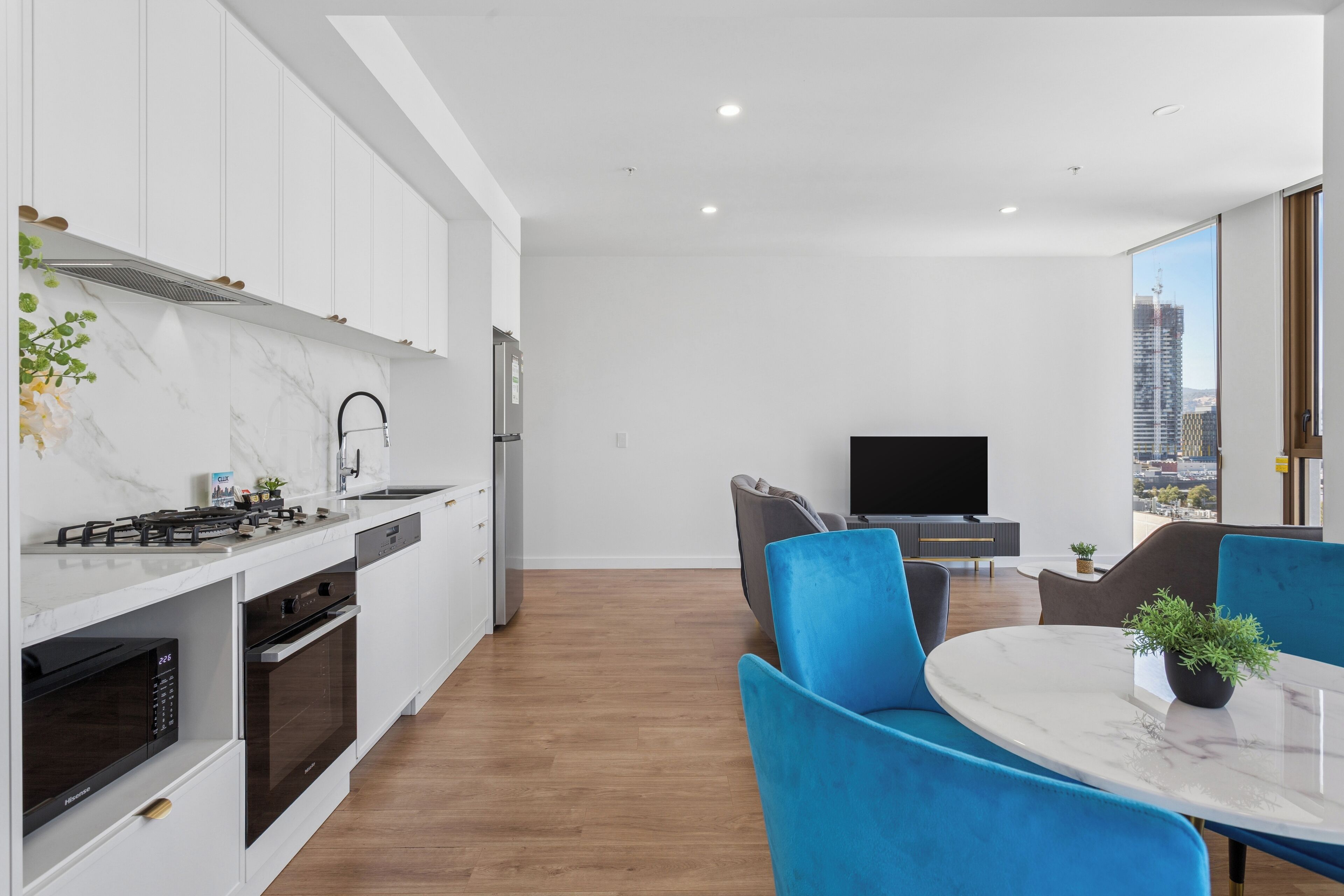 A modern kitchen with white cabinets, a marble countertop, and a built-in oven. There is a dining area with blue chairs and a table, and a TV in the background.