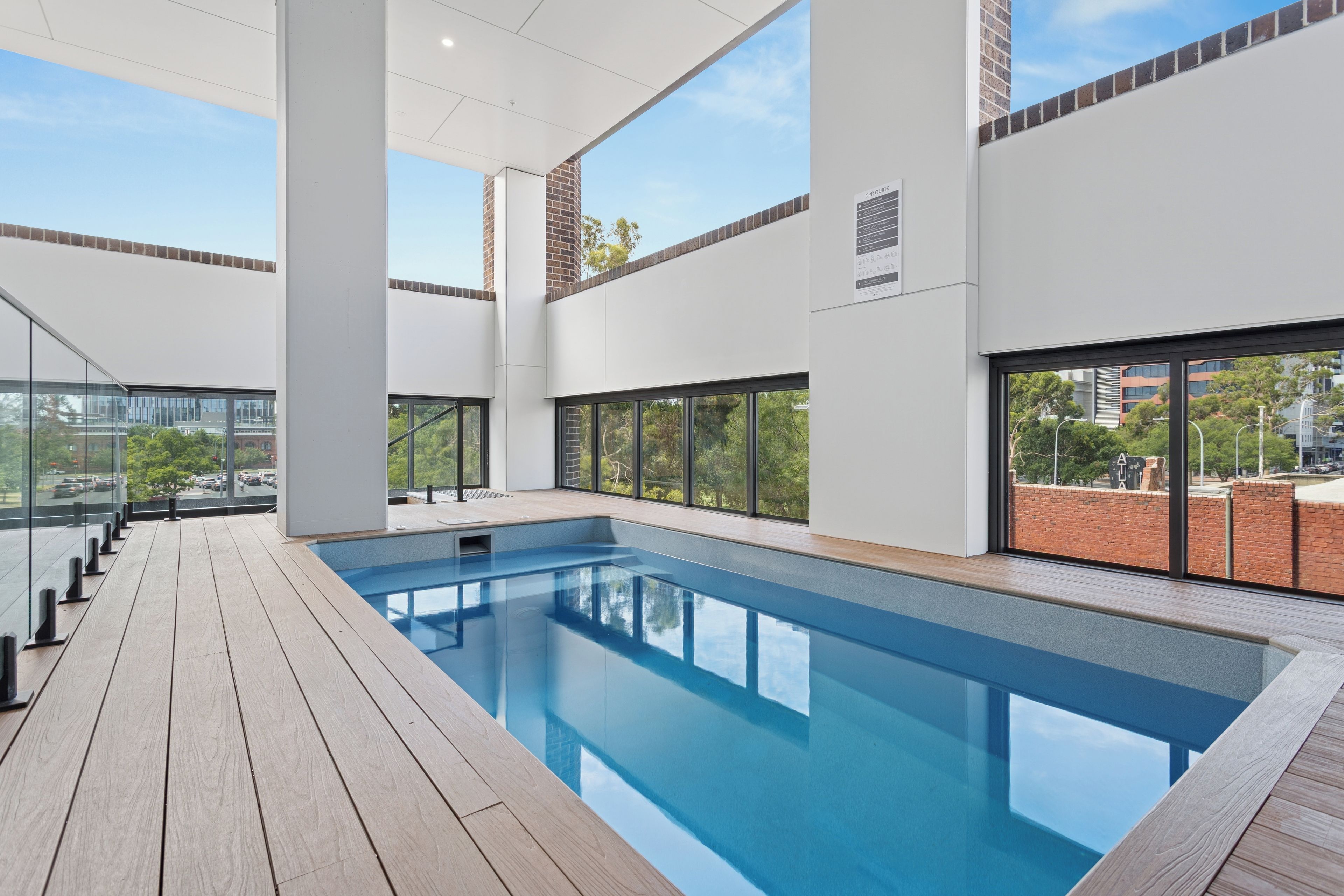 Une piscine intérieure moderne avec des parois en verre transparent, une terrasse en bois et une vue sur le paysage urbain.