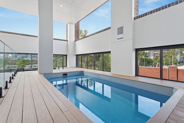 Una piscina interior moderna con paredes de vidrio transparente, una terraza de madera y vistas a la ciudad.