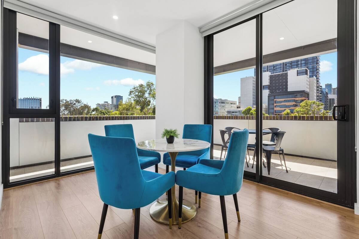 A modern room with a round table and blue chairs, large windows with a city view, and a balcony with outdoor seating.