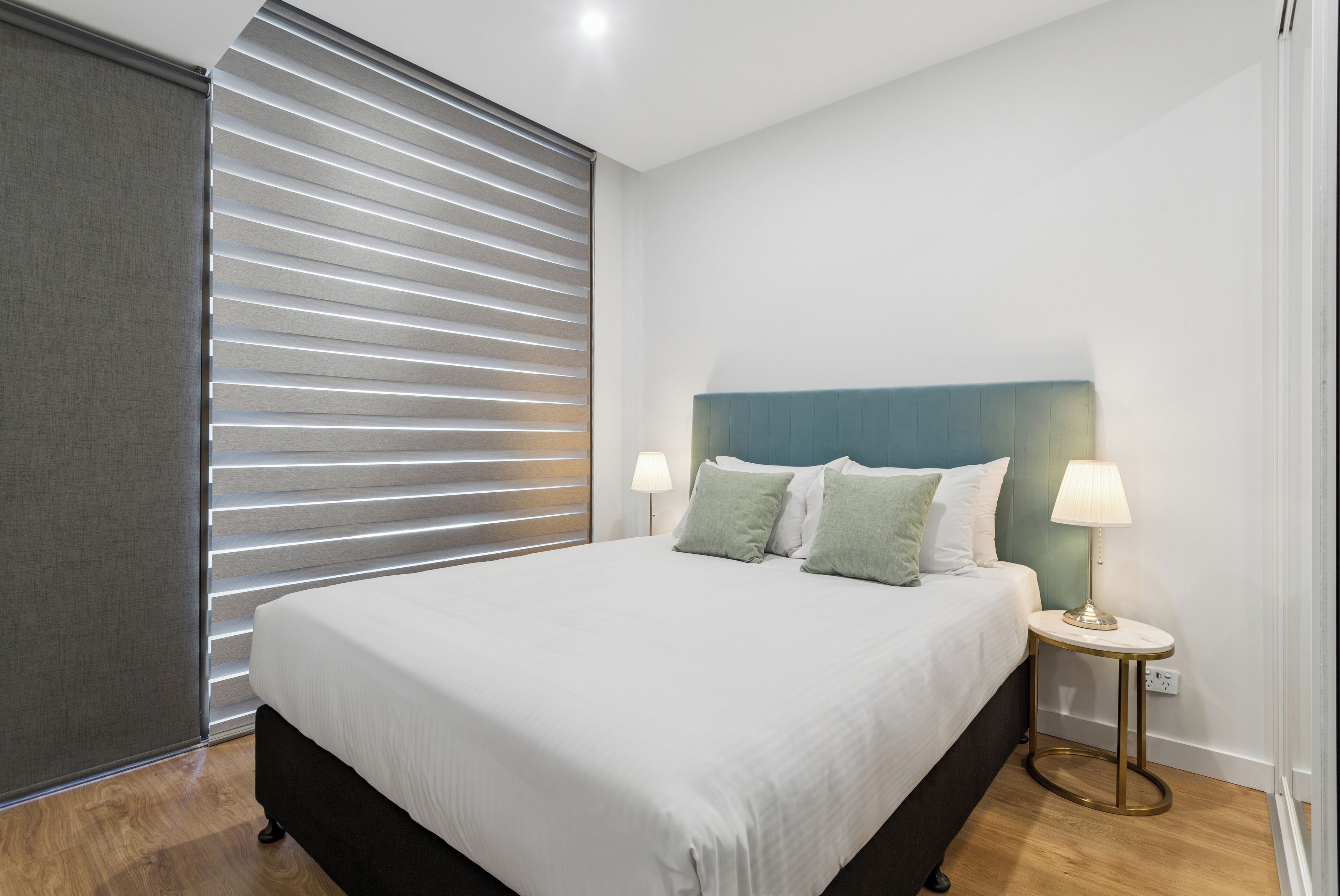 A neatly made bed with a white duvet and two pillows, a bedside table with a lamp, and a window with horizontal blinds.