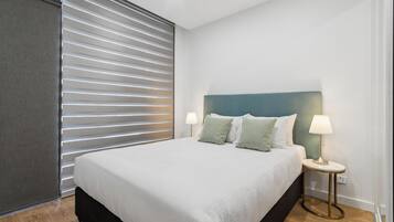 A neatly made bed with a white duvet and two pillows, a bedside table with a lamp, and a window with horizontal blinds.