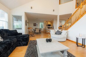 Un vaste espace de vie comprenant un canapé noir, un fauteuil blanc et une table basse. On aperçoit un escalier avec des rampes en bois à l’arrière-plan.