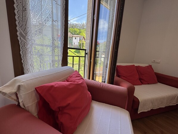 A room with a red sofa and a white sofa, a lace curtain, and a view of a green landscape through a window.
