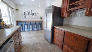 A kitchen with a stainless steel refrigerator, wooden cabinets, a bar stool area, and a 'gather' sign on the wall. - Entire House Between Zion & Bryce w/ Hot Tub! (Orderville)