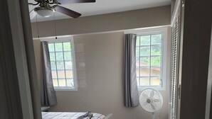 Ein Schlafzimmer mit einem Bett, Deckenventilator, Standventilator und einem Fenster mit Vorhängen.