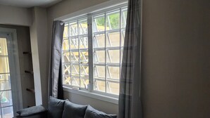 Ein Zimmer mit einer grauen Couch, einem kleinen Hocker, einem Fenster mit Metallgitter und einer Tür mit Glasfeldern.