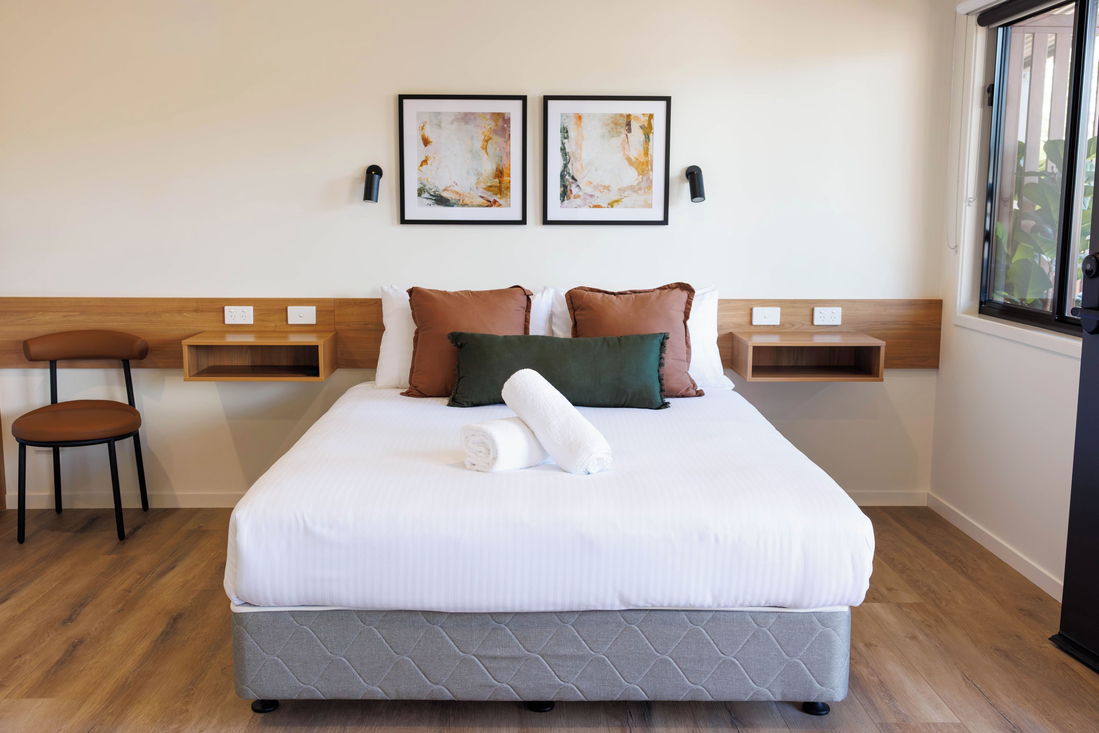 A neatly made bed with a quilted headboard, two bedside tables, a chair, and two abstract paintings on the wall.