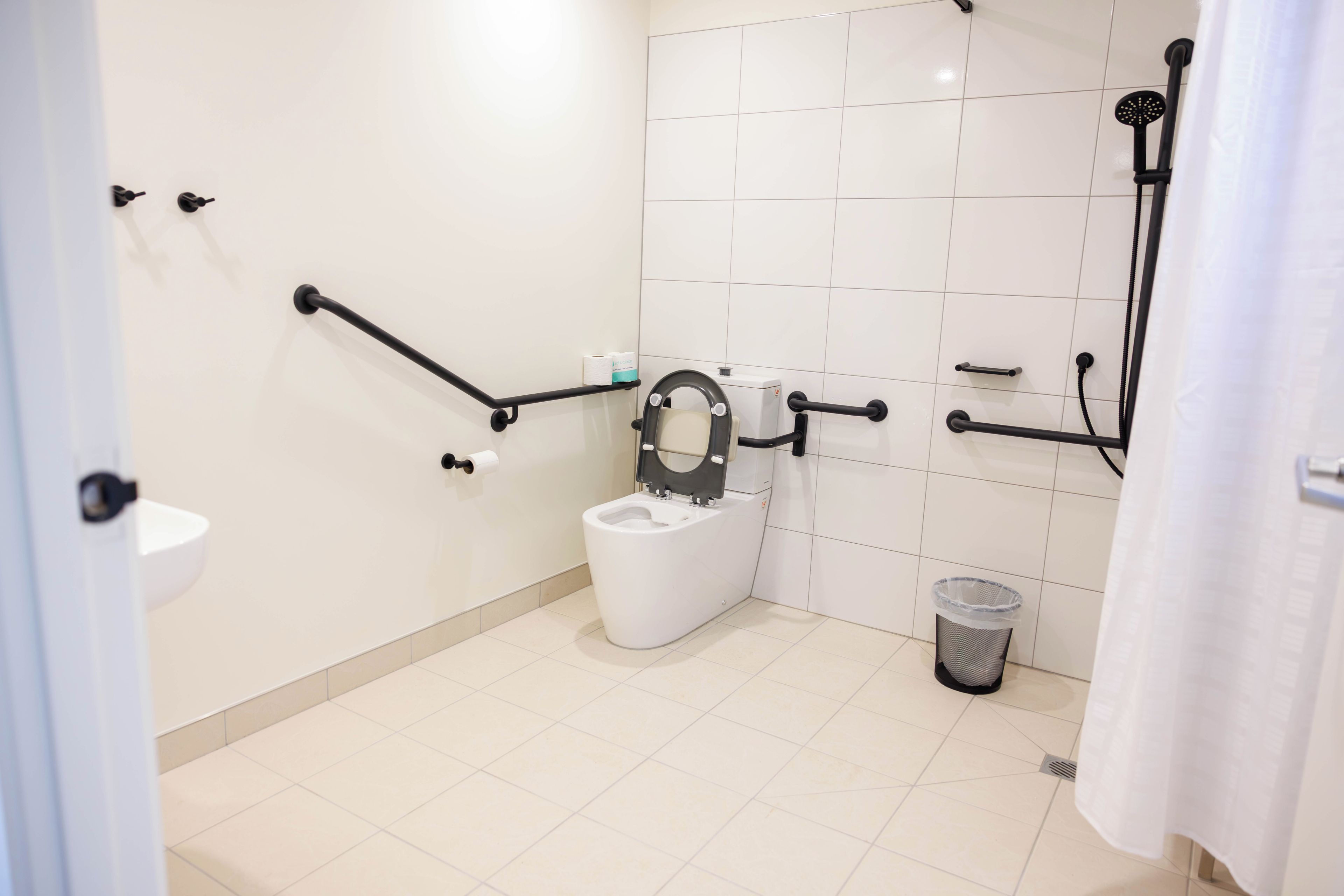 A modern bathroom with a toilet, handrails, a shower, and a trash bin.