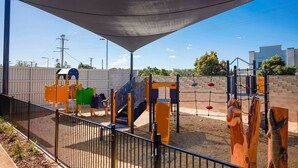 A playground with various play structures, including slides and climbing areas, surrounded by a fence and a shaded canopy. - Coomera Lodge Hotel (Oxenford)