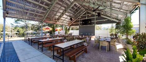 An outdoor patio with wooden tables and benches, a covered area with a metal roof, and potted plants.