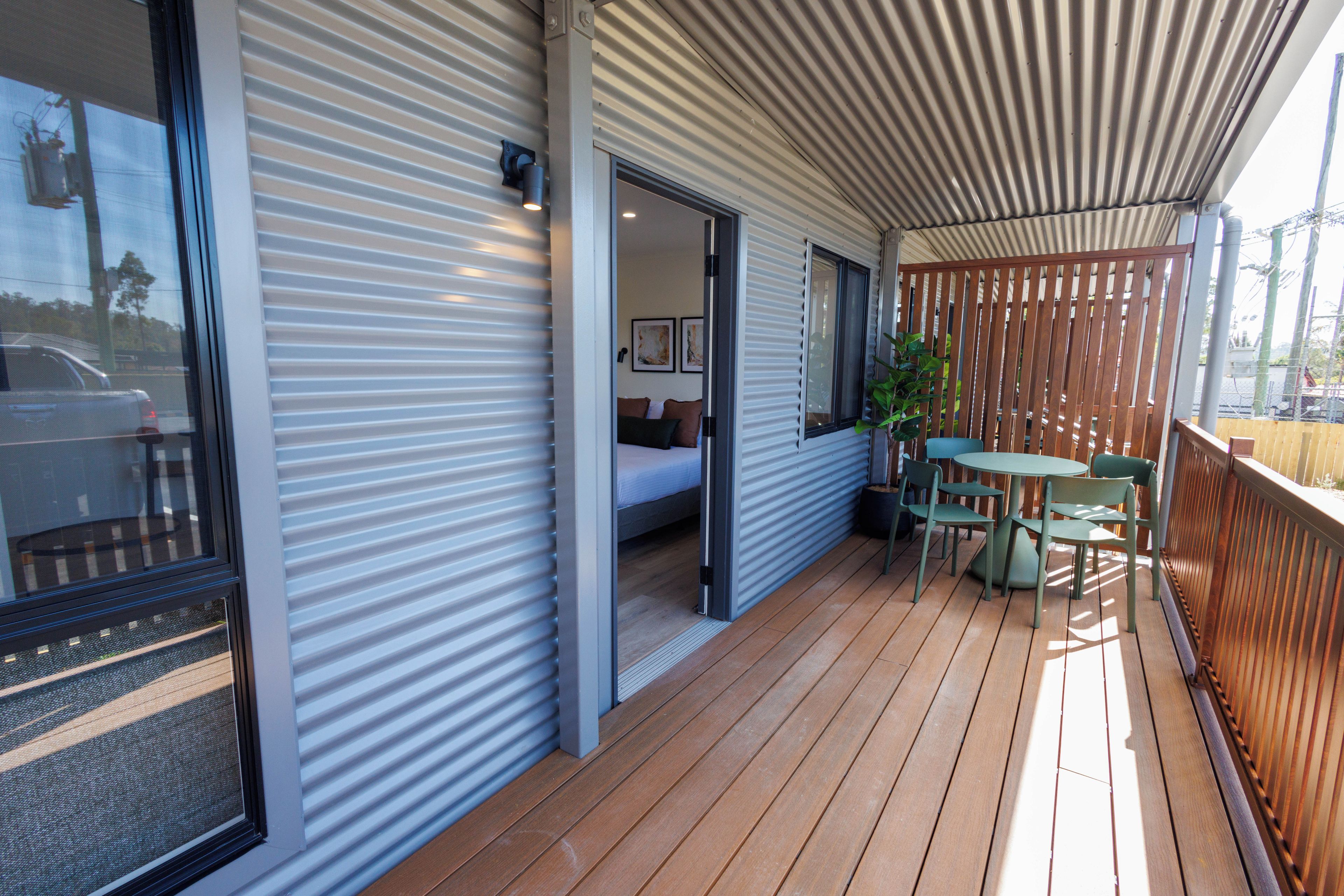 A covered outdoor area with a wooden deck, a seating arrangement, and a door leading to a room with a bed and a painting.
