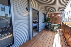 Un espacio al aire libre techado con piso de madera, una zona de estar y una puerta que da a una habitación con cama y un cuadro.