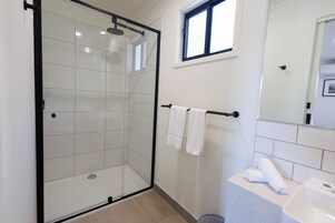 Egy modern fürőszoba üvegfalas zuhanyzóval, két törölközővel ellátott törölközőtartóval és tükörrel rendelkező mosdókagylóval.