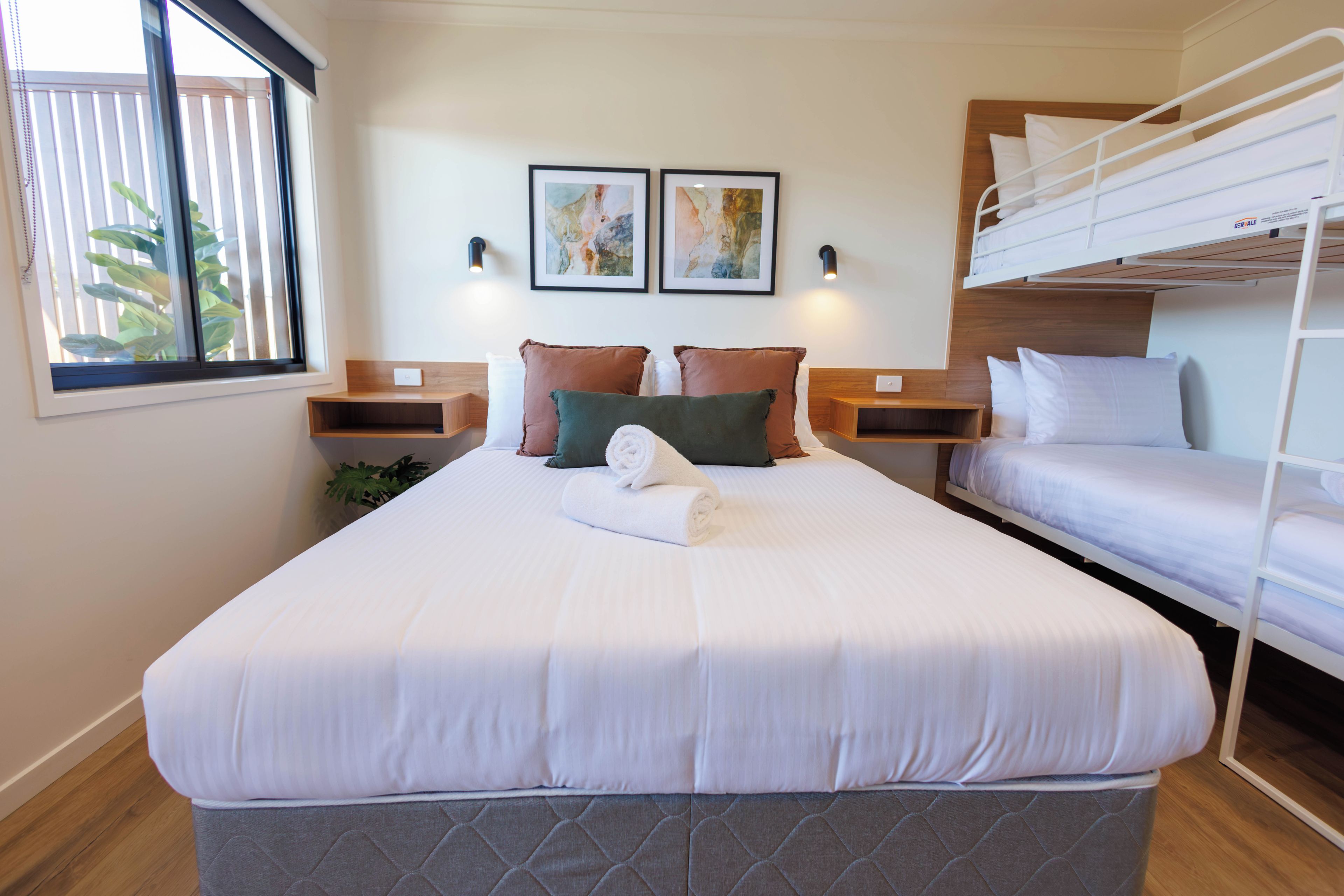 A hotel room with a single bed, a bunk bed, a bedside table, a wall-mounted light, and a window with blinds.