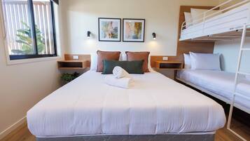 A hotel room with a single bed, a bunk bed, a bedside table, a wall-mounted light, and a window with blinds.