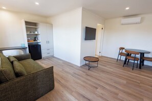 A modern living room with a sofa, a dining table with chairs, a kitchen area with cabinets, and a flat-screen TV mounted on the wall. - Coomera Lodge Hotel (Oxenford)