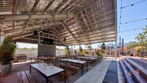 Eine Terrasse im Freien mit Holztischen und -bänken, ein überdachter Bereich mit Metallveranda und ein Betonweg.