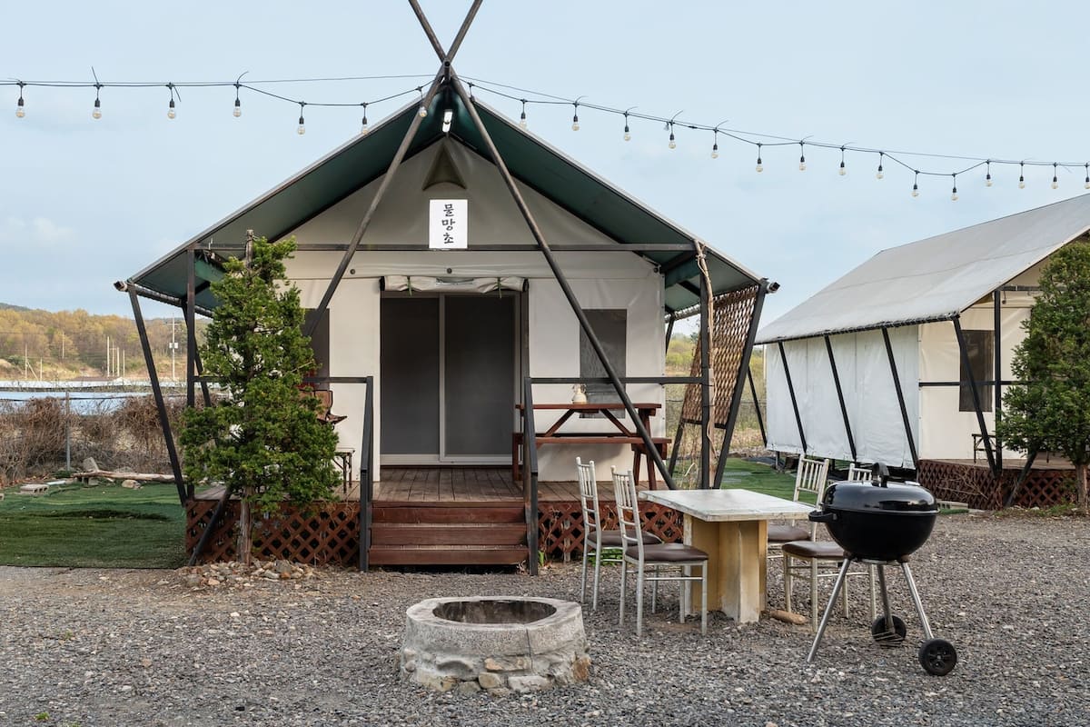 A small, single-story tent with a door, a table with chairs, a barbecue grill, and a fire pit.