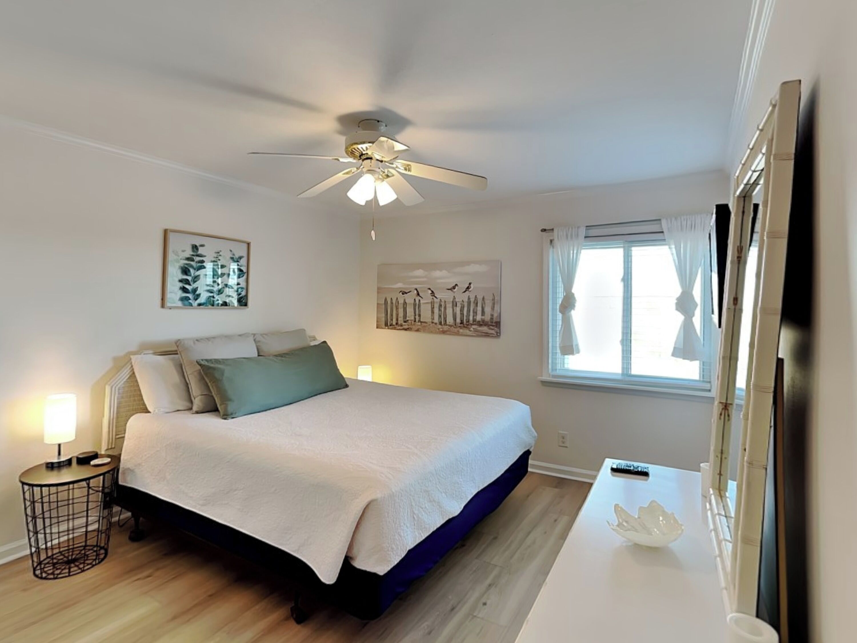 A bedroom with a bed, a ceiling fan, a window with curtains, a bedside table with a lamp, and wall art.
