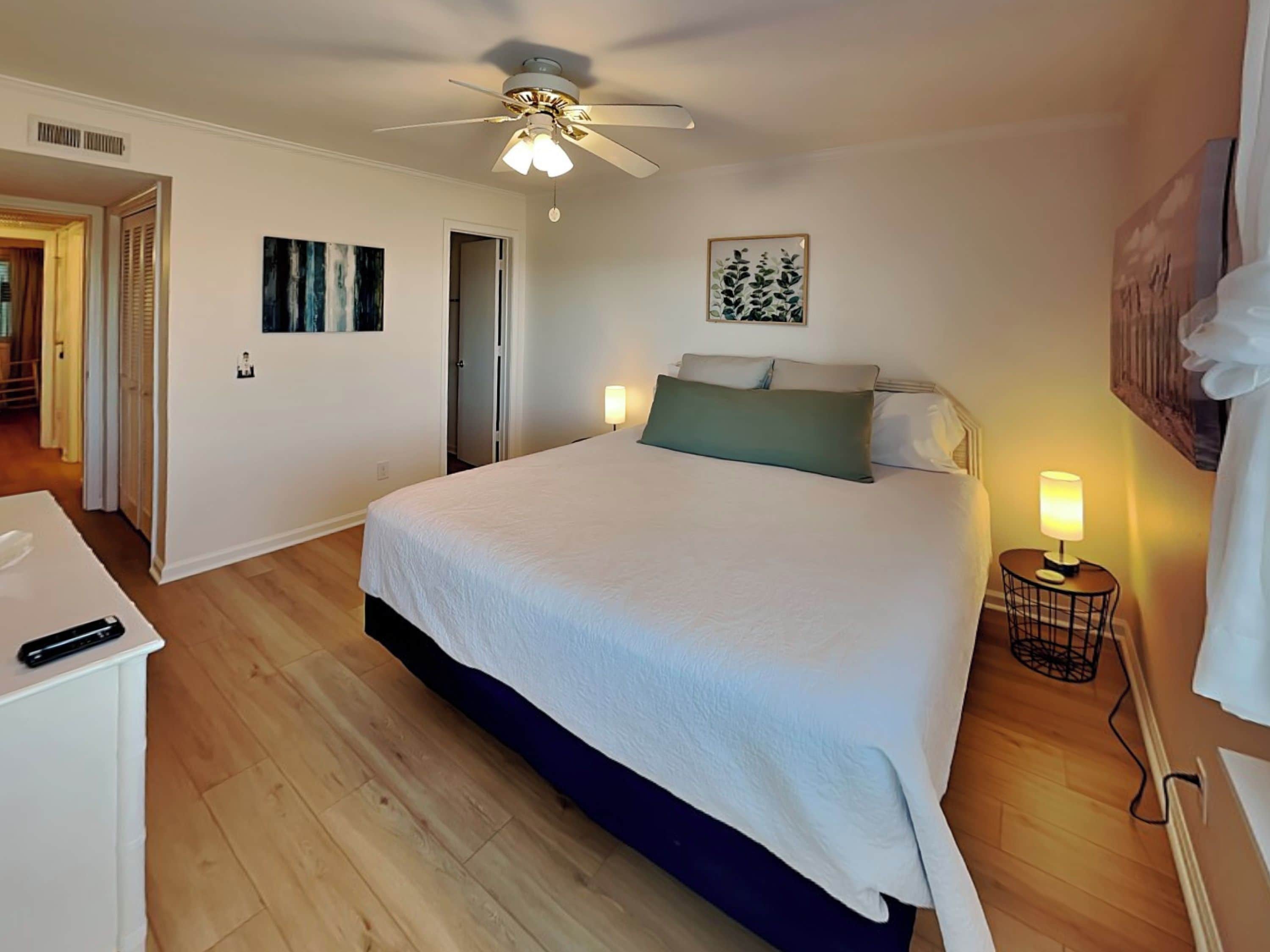 A bedroom with a bed, bedside lamps, a ceiling fan, and a door leading to another room.