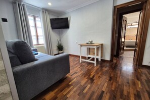 A room with a grey sofa, wooden flooring, a TV mounted on the wall, and a small table with a plant.