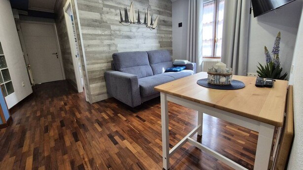 A room with a grey sofa, a wooden table, a wall-mounted TV, and a window with curtains. - 1a152 Apartamentos el Veleru Popa (Gijón)