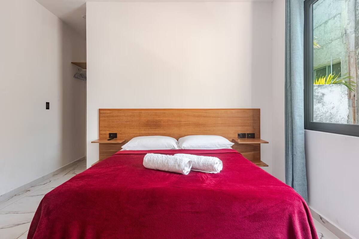 A hotel room with a wooden headboard, two white pillows, a red blanket, and a rolled white towel on the bed.
