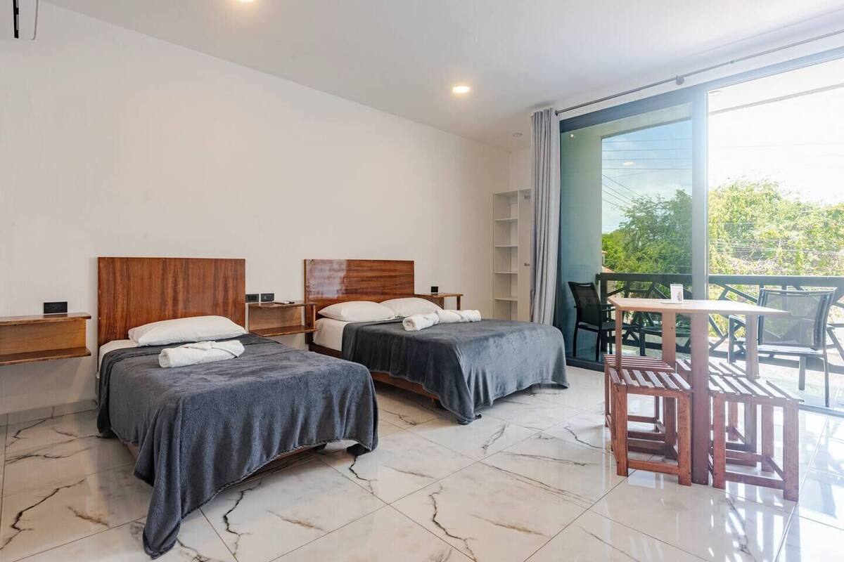 A modern hotel room with two beds, a balcony with a table and chairs, and a large window providing a view of greenery.