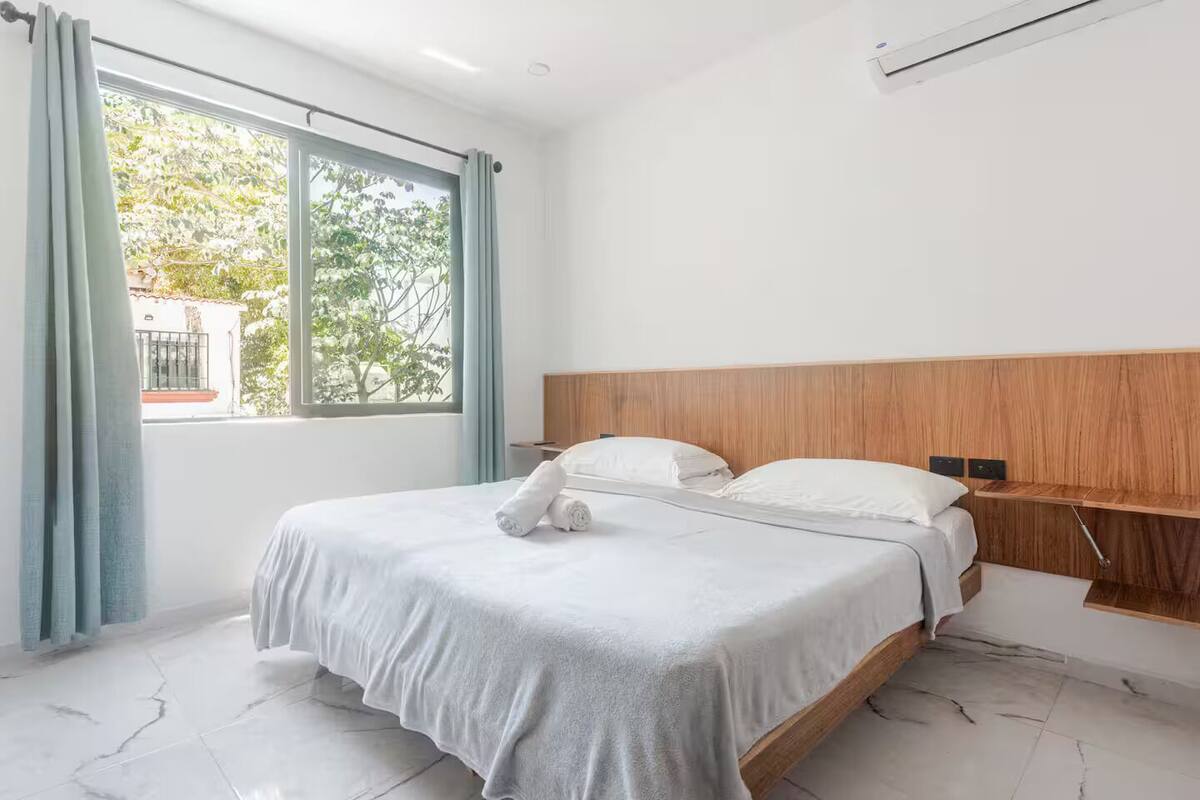 A modern bedroom with a large bed, a wooden headboard, and a view of greenery outside.