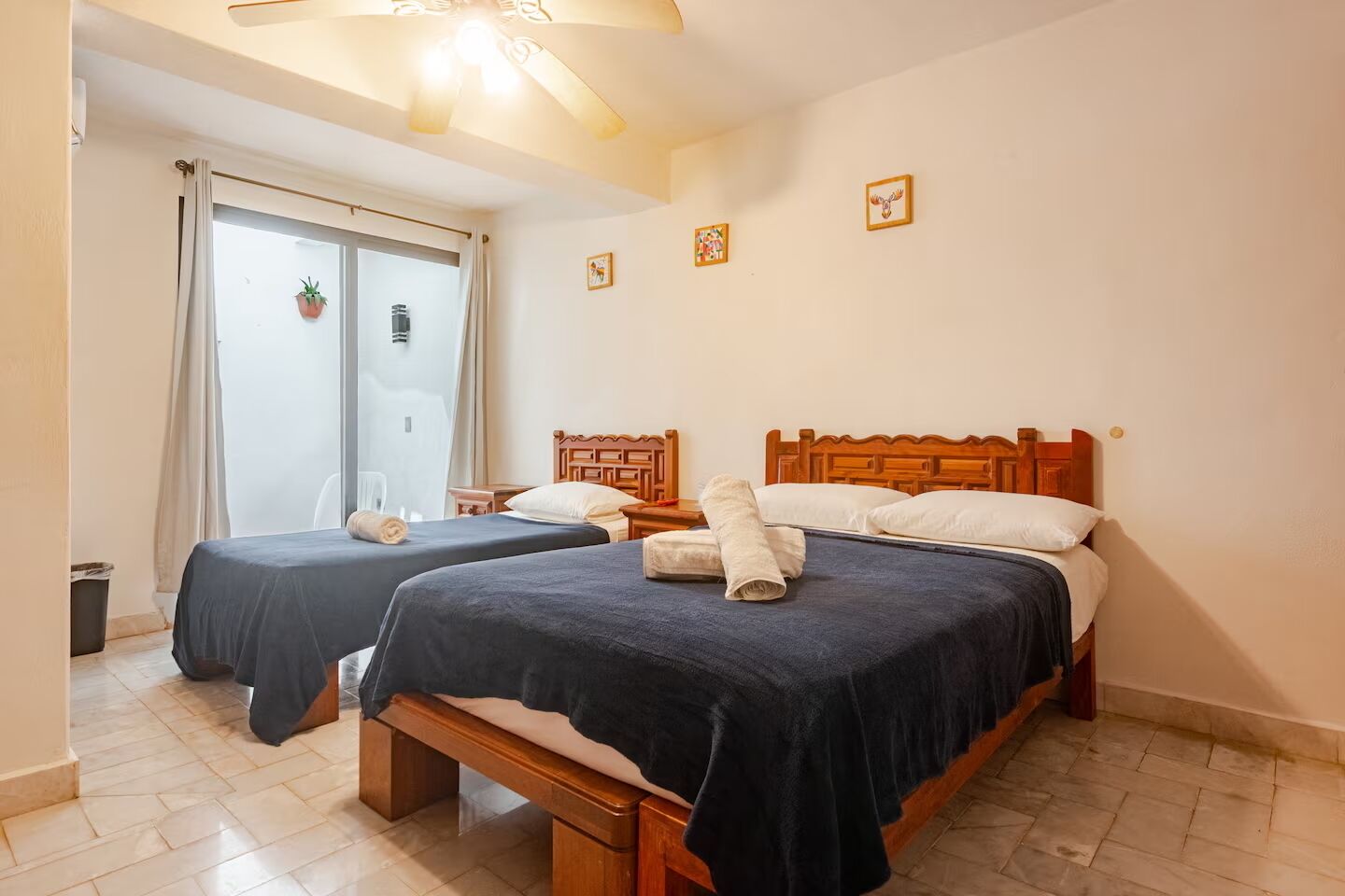 Quarto de hotel com duas camas, um ventilador de teto, janela com cortinas e uma pequena planta em vaso.
