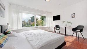 Une chambre d’hôtel moderne avec un grand lit, une télévision, une petite table, deux chaises et une fenêtre donnant sur un paysage verdoyant.
