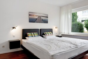 A modern bedroom with a large bed, two bedside tables, a wall-mounted artwork, and a window with a view. - Lake Constance Bungalow – Apartment 'Bungalow' with Private Terrace, Shared Garden & Wi-Fi (Immenstaad am Bodensee)