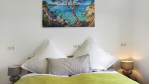 A bedroom with a bed, pillows, a nightstand, a lamp, and a wall-mounted painting of a coastal landscape. - Herkules Suite Apartment in Kassel (Kassel)