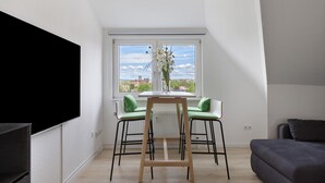 A modern living room with a TV, a dining table with chairs, a sofa, and a window with a view. - Herkules Suite Apartment in Kassel (Kassel)